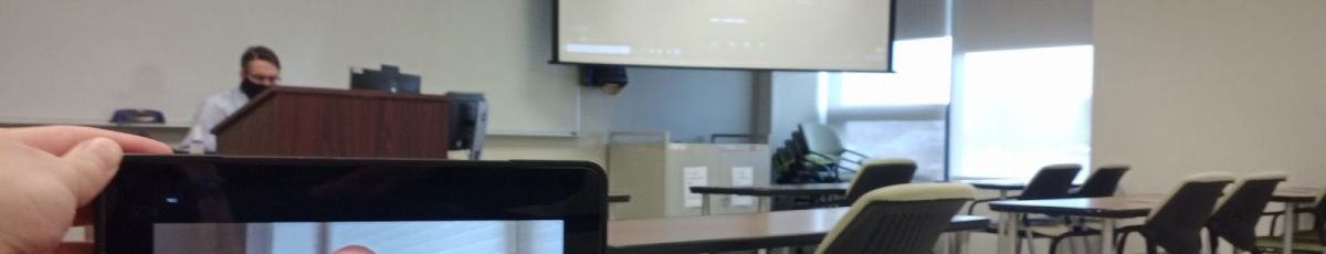 Image of Flex classroom from student perspective with device in foreground and teacher, projector screen in background