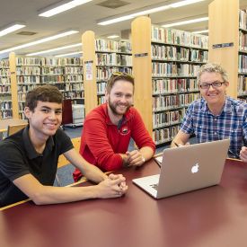 Two male students working with faculty on laptop in library