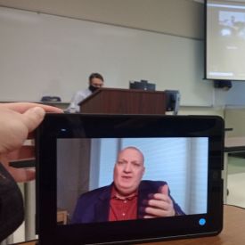 Image of Flex classroom from student perspective with device in foreground and teacher, projector screen in background