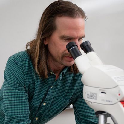 Dr. Evan Thomas - Assistant Professor of Biology looking through microscope (square)