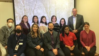 FET group photo with twelve members in front of a screen in a flex room. Five people are sitting down and twelve are standing up behind them.