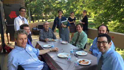 Casual group photo of ten FET members outside, with six sitting at a picnic table and four standing behind them. Several plates are on the table.